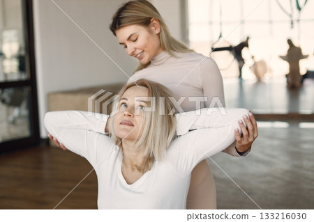 Blonde woman doing yoga with personal trainer. Yoga trainer assisting woman while doing exercises. Blonde woman wearing white clothes and trainer wearing beige costume. 133216030