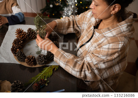 Family Decorating Christmas Wreath With Pine Cones And Greenery At Home 133216086