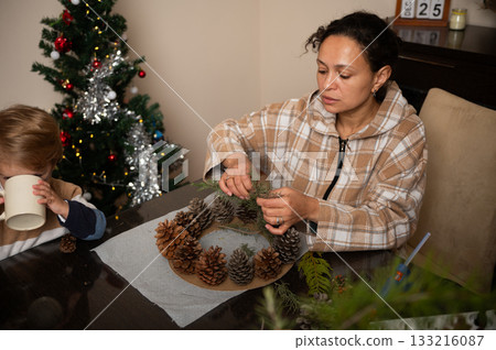 Family Decorating Christmas Wreath With Pinecones And Evergreen Branches At Cozy Home Table Family Decorating Christmas Wreath With Pinecones And Evergreen Branches At Cozy Home Table 133216087