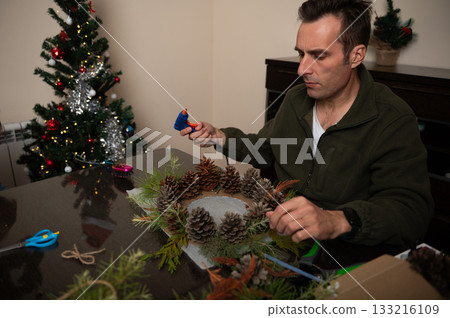 Man Crafts Christmas Wreath With Pinecones At Home For Cozy Holiday Decor Man Crafts Christmas Wreath With Pinecones At Home For Cozy Holiday Decor 133216109