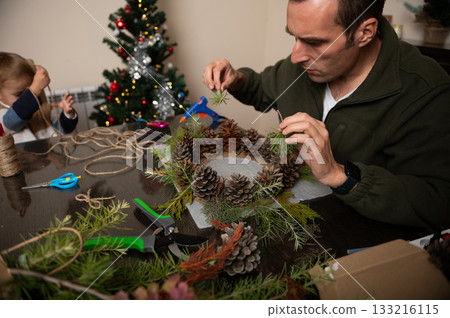 Family Decorates Christmas Wreath With Pinecones And Greenery In Cozy Home Setting Family Decorates Christmas Wreath With Pinecones And Greenery In Cozy Home Setting 133216115