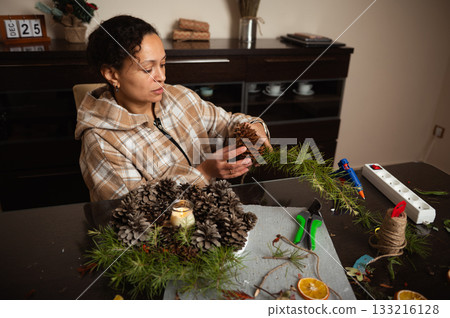 Woman Crafting Christmas Pine Cone Decorations at Home with Greenery and Candles Woman Crafting Christmas Pine Cone Decorations at Home with Greenery and Candles 133216128