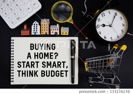 Notepad with the phrase "Buying A Home? Start Smart, Think Budget" surrounded by a pen, mini trolley shopping cart, magnifying glass, clock, and house model 133216238