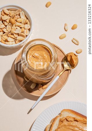 Peanut butter in a glass jar on a light background with scattered nuts, spoon and toast. Peanut butter in a glass jar on a light background with scattered nuts, spoon and toast. 133216324