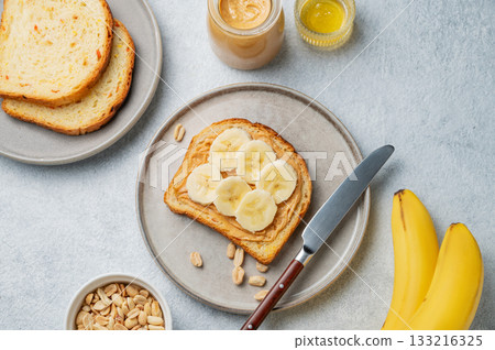 Peanut butter toast and banana in a plate on a light background with nuts, knife and honey. 133216325