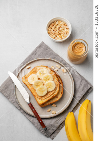Peanut butter toast and banana in a plate on a light background with nuts, jar of spread and knife. Peanut butter toast and banana in a plate on a light background with nuts, jar of spread and knife. 133216326