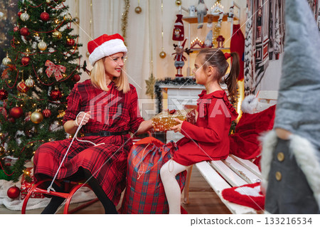 Mother wearing Santa hat giving wrapped present to young daughter Mother wearing Santa hat giving wrapped present to young daughter 133216534