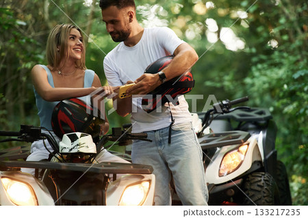 Holding yellow smartphone, showing something. Man and woman are on ATV outdoors 133217235