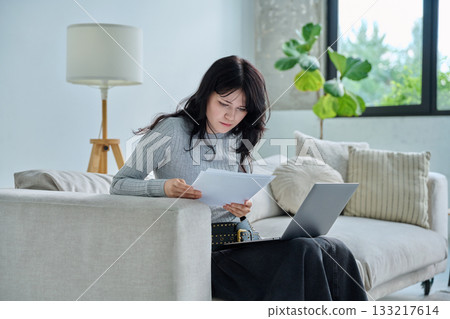 Young serious student girl sitting at home on couch with laptop, reading papers 133217614
