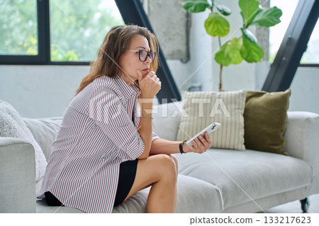 Sad serious middle aged woman with smartphone sitting on sofa 133217623