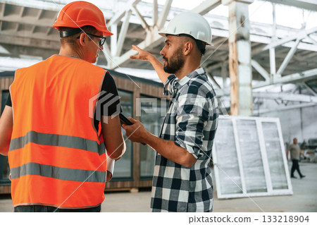 Builder is talking with manager or architect. Two workers are in the industrial wooden warehouse 133218904