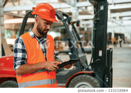 Forklift is behind, digital tablet in hands. Industrial worker in wooden warehouse Forklift is behind, digital tablet in hands. Industrial worker in wooden warehouse 133218964