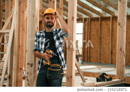 Orange colored drill and hardhat in hands. Industrial worker in wooden warehouse Orange colored drill and hardhat in hands. Industrial worker in wooden warehouse 133218971