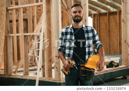 Building the house, holding hard hat and drill. Industrial worker in wooden warehouse Building the house, holding hard hat and drill. Industrial worker in wooden warehouse 133218974