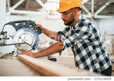 By the table, using blade, cutting the fresh wood. Industrial worker in warehouse 133219009