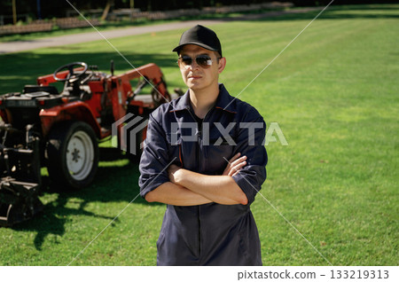 Standing with arms crossed. Man is with utility tractor with grass cutter and aerator equipment on the field 133219313