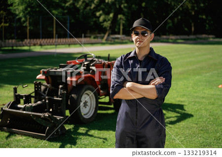 Standing with arms crossed. Man is with utility tractor with grass cutter and aerator equipment on the field 133219314