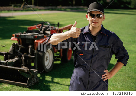 Shows thumb up. Man is with utility tractor with grass cutter and aerator equipment on the field Shows thumb up. Man is with utility tractor with grass cutter and aerator equipment on the field 133219316