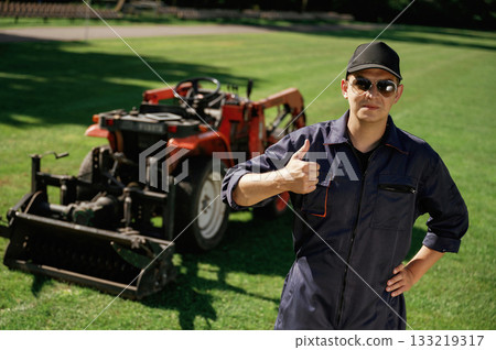 Shows thumb up. Man is with utility tractor with grass cutter and aerator equipment on the field 133219317