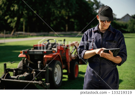 Black digital tablet in hands. Man is with utility tractor with grass cutter and aerator equipment on the field Black digital tablet in hands. Man is with utility tractor with grass cutter and aerator equipment on the field 133219319