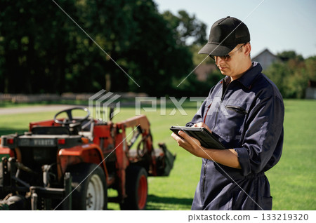 Black digital tablet in hands. Man is with utility tractor with grass cutter and aerator equipment on the field Black digital tablet in hands. Man is with utility tractor with grass cutter and aerator equipment on the field 133219320