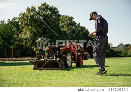Rear, back view, walking. Man is with utility tractor with grass cutter and aerator equipment on the field 133219324