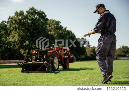 Rear, back view, walking. Man is with utility tractor with grass cutter and aerator equipment on the field 133219325