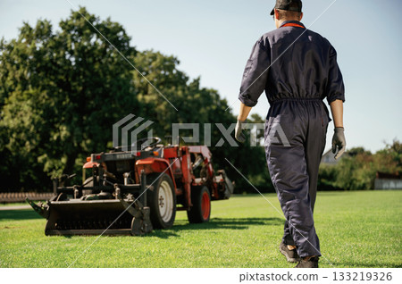 Rear, back view, walking. Man is with utility tractor with grass cutter and aerator equipment on the field 133219326