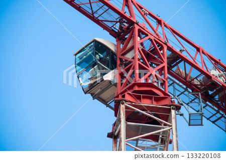 Close-up view of Control Cabin of Red-and-white Tower Crane on Construction Site against Blue Sky on Sunny Day. Fasteners of Crane and Metal Parts are covered with Rust Close-up view of Control Cabin of Red-and-white Tower Crane on Construction Site against Blue Sky on Sunny Day. Fasteners of Crane and Metal Parts are covered with Rust 133220108