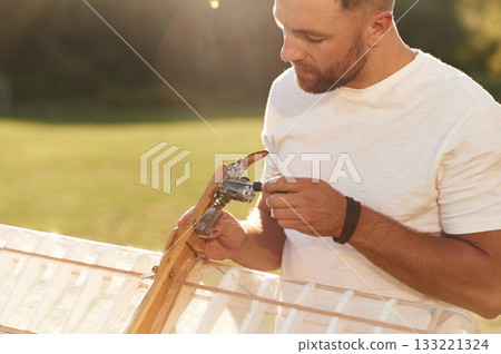 Made of wood. Man is on the field fixing the toy plane Made of wood. Man is on the field fixing the toy plane 133221324