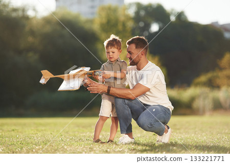 Looking at it, sitting. Father and son are on the field together with toy plane Looking at it, sitting. Father and son are on the field together with toy plane 133221771