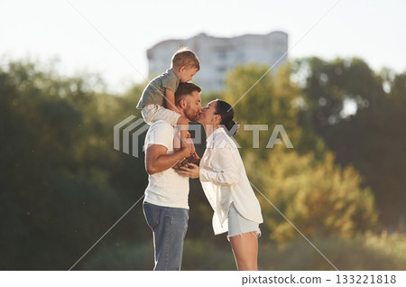 Kid is sitting on the shoulders. Mother and father are with son outdoors on the field 133221818