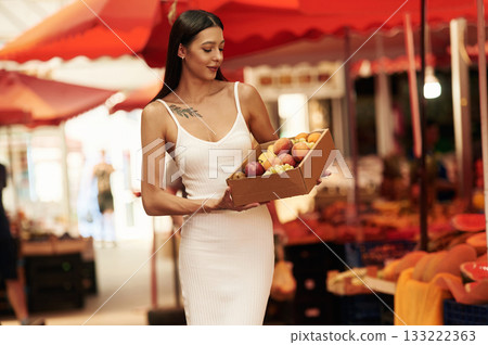 Grapes, peaches, and other fruits in a box. Young woman is on the vegetable market or bazaar 133222363