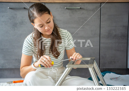 Smiling young woman assembling a chair with tools on the floor. Concept of joy and satisfaction from independent home projects. Smiling young woman assembling a chair with tools on the floor. Concept of joy and satisfaction from independent home projects. 133222385