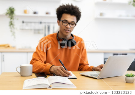 A young man with glasses writes in a notebook while working on a laptop at home, with a cup of coffee and a plant on the table. 133223123