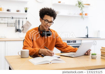 A young Black man wearing glasses and headphones works on a laptop while taking notes in a kitchen setting. 133223125