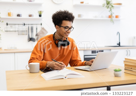 A young man with glasses works on a laptop while taking notes in a kitchen setting, smiling. 133223127