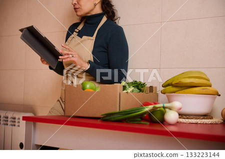 Woman in Apron Using Tablet While Reviewing Fresh Groceries in a Kitchen Setup 133223144