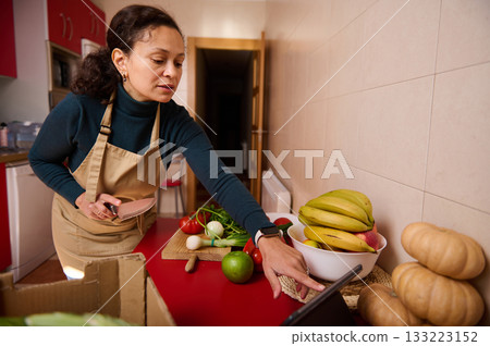 Woman In Apron Reaches For Tablet While Preparing Fresh Fruit And Vegetables In Modern Kitchen Woman In Apron Reaches For Tablet While Preparing Fresh Fruit And Vegetables In Modern Kitchen 133223152