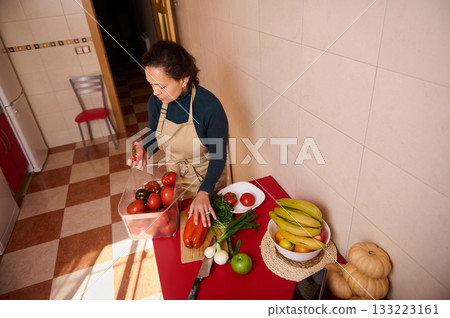 Woman In Apron Preparing Fresh Produce With Tomatoes, Bananas, And Pumpkins In Kitchen Woman In Apron Preparing Fresh Produce With Tomatoes, Bananas, And Pumpkins In Kitchen 133223161