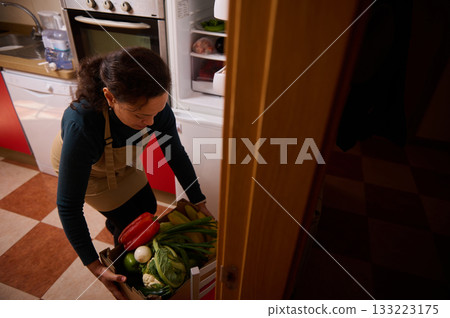 Woman In Apron Carries Box Of Fresh Vegetables In Kitchen For Healthy Meal Prep Woman In Apron Carries Box Of Fresh Vegetables In Kitchen For Healthy Meal Prep 133223175