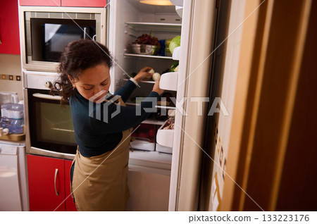 Woman in Apron Organizing Fresh Food in Open Refrigerator in Modern Kitchen Woman in Apron Organizing Fresh Food in Open Refrigerator in Modern Kitchen 133223176