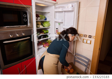 Woman Organizing Fresh Produce From Open Refrigerator In Kitchen Woman Organizing Fresh Produce From Open Refrigerator In Kitchen 133223183
