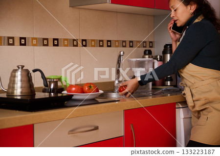 Woman Washing Tomato At Kitchen Sink While Talking On The Phone In A Red And Beige Kitchen Woman Washing Tomato At Kitchen Sink While Talking On The Phone In A Red And Beige Kitchen 133223187