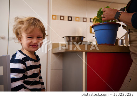 Happy Toddler In Striped Shirt Smiles At Camera In Kitchen While Parent Prepares Food 133223222