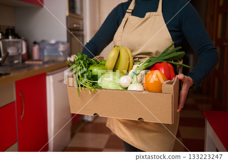 Person In Apron Holding Box Of Fresh Groceries In Cozy Kitchen Person In Apron Holding Box Of Fresh Groceries In Cozy Kitchen 133223247