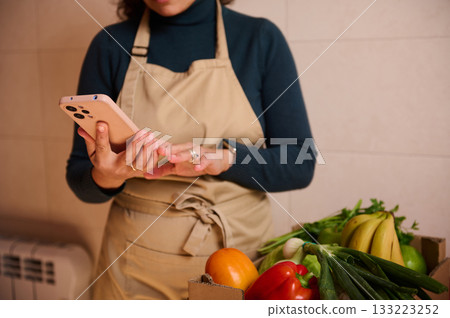 Woman In Apron Using Phone By Fresh Produce Box At Home Market Woman In Apron Using Phone By Fresh Produce Box At Home Market 133223252