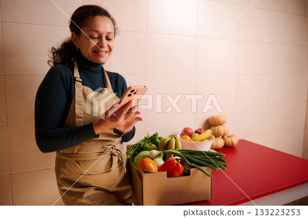 Smiling Woman With Apron Checks Her Phone Beside Fresh Produce Box On Red Counter Smiling Woman With Apron Checks Her Phone Beside Fresh Produce Box On Red Counter 133223253