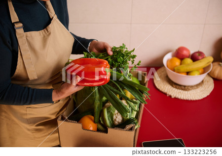 Person In Kitchen Apron Holding Red Pepper Over Box Of Fresh Produce Person In Kitchen Apron Holding Red Pepper Over Box Of Fresh Produce 133223256