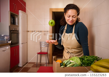 Woman In Apron Tossing Apple While Preparing Fresh Vegetables In Cozy Kitchen Woman In Apron Tossing Apple While Preparing Fresh Vegetables In Cozy Kitchen 133223263